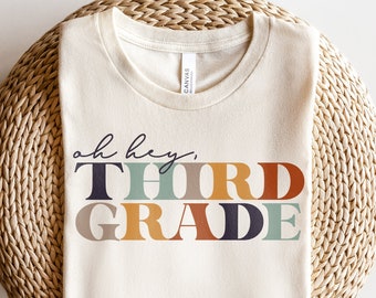 May include: A beige t-shirt with the text "oh hey, THIRD GRADE" in colourful block letters. The letters are in shades of green, orange, brown, and blue.