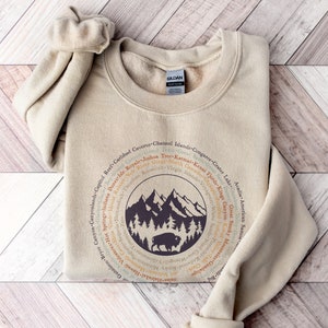 May include: A sand-colored crewneck sweatshirt featuring a circular graphic design. The center displays a dark purple illustration of mountains, pine trees, and a bison. Surrounding the central image are the names of various national parks.