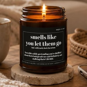May include: A lit candle in a dark amber glass jar with a black label featuring white text that reads: smells like you let them go, but still track their location. burn this while pretending you're thrilled about their independence and definitely not stalking their Life360.