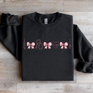 May include: Black sweatshirt with a pink medical-themed design. The design includes a syringe, bows, a stethoscope, a heart with a heartbeat line, and a thermometer. The sweatshirt is made of a soft material.