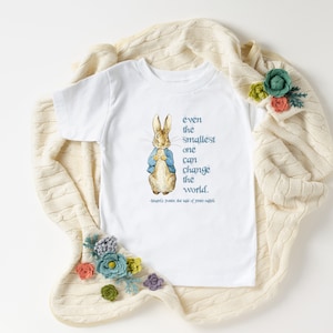 May include: White toddler t-shirt featuring a watercolor illustration of Peter Rabbit wearing a blue jacket. The shirt has the quote "even the smallest one can change the world" in teal text. The quote is attributed to Beatrix Potter.