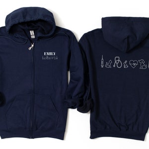 Peut inclure: Sweat-shirt à capuche bleu marine zippé avec le nom "Emily" et un cœur avec un stéthoscope, une seringue, un chien, un chat et un thermomètre au dos.