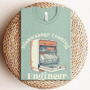 May include: A light green t-shirt with a retro-style illustration of a dishwasher with plates inside. The text "Dishwasher Loading Engineer" is written in a curved font around the dishwasher.