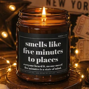 May include: A lit brown glass jar candle with a black label that reads "smells like five minutes to places." The candle sits on a wooden coaster, surrounded by fairy lights and theater tickets. The candle is a maple and hen 100% natural soy wax blend.