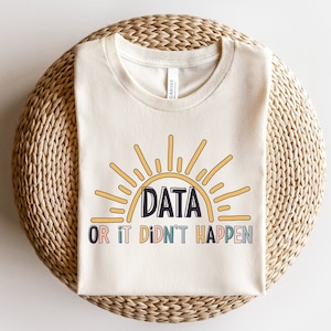 May include: A cream-coloured t-shirt featuring the text "DATA OR IT DIDN'T HAPPEN" within a colourful sun design. The shirt is folded on a woven, circular surface.