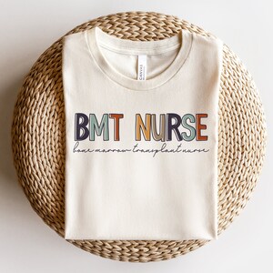 May include: Cream-colored t-shirt with the text "BMT NURSE" in colorful block letters. Below the text, the words "bone marrow transplant nurse" are written in a cursive font. The shirt is folded and resting on a woven, circular surface.
