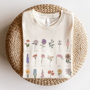 Peut inclure: Un t-shirt crème avec un motif de 20 fleurs différentes et leurs noms imprimés sur le devant. Les fleurs sont dessinées dans un style vintage avec des détails délicats.