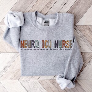 May include: A gray sweatshirt with the text "NEURO ICU NURSE" in colorful letters. The text "neuro intensive care unit" is written in smaller letters below the main text.