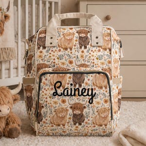 May include: A floral-patterned diaper bag featuring illustrations of Highland cows. The bag has multiple pockets and a top handle. The name "Lainey" is written in cursive on the front. A stuffed Highland cow and a baby blanket are also in the image.