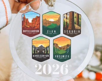 May include: A clear, circular glass ornament featuring five hexagonal illustrations of national parks: Bryce Canyon, Zion, Sequoia, Kings Canyon, and Yosemite. The year 2026 is printed in white at the bottom. A gold ribbon is attached to the top.