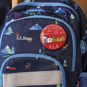 May include: Navy blue backpack with a mountain and tree pattern, featuring a reflective strip and blue trim. A red button with allergy information is attached. A water bottle with the text "EXPLORE MORE" is visible.