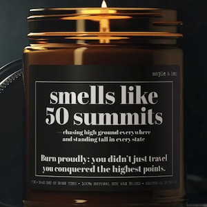 May include: A lit candle in a brown glass jar with a black label that reads "smells like 50 summits." The jar has a gold lid and is sitting on a dark surface. The text on the label includes the phrases "chasing high ground" and "Burn proudly."