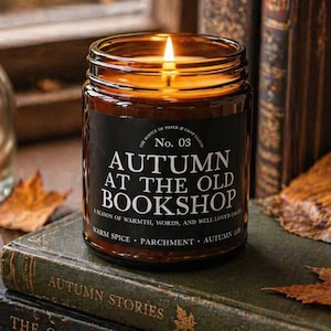 May include: A lit candle in a glass jar with a black label that reads "AUTUMN AT THE OLD BOOKSHOP" sits atop old books. The scene includes dried leaves, a cup, and a vase with dried flowers, creating a cozy, autumnal ambiance.