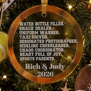 May include: A round, transparent acrylic Christmas ornament with a gold-toned border and a gold ribbon hanger. The text reads: WATER BOTTLE FILLER. SNACK DEALER. UNIFORM WASHER. TAXI DRIVER. DESIGNATED PHOTOGRAPHER. SIDELINE CHEERLEADER. CHAOS COORDINATOR. HEART FULL OF JOY. SPORTS PARENTS. Rich & Judy 2026.