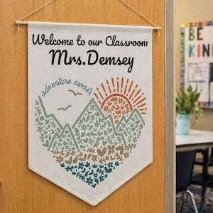 May include: A classroom banner reading "Welcome to our Classroom Mrs. Demsey" and "adventure awaits". The design includes mountains and a sun in green, brown, and orange. It is suspended from a wooden door.