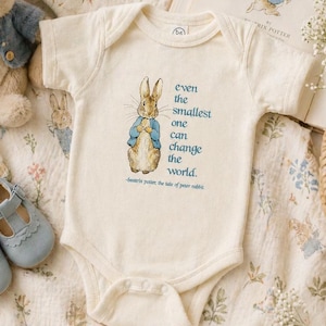 May include: A cream-colored baby onesie featuring a Peter Rabbit illustration and the quote, "even the smallest one can change the world." The onesie is surrounded by a plush rabbit, baby shoes, and a book titled "The Tale of Peter Rabbit."