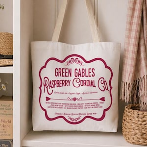 May include: A white tote bag with cream-colored handles hangs on a hook. The bag features a burgundy-colored graphic with the text "Green Gables Raspberry Cordial Co." and additional text. A straw hat and books are visible on a shelf.