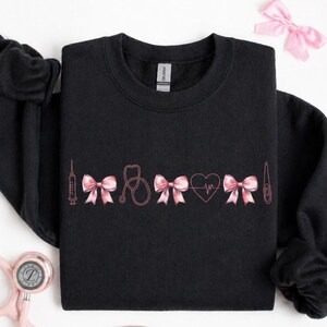 May include: Black sweatshirt with pink medical-themed embroidery. The design includes a syringe, bows, a stethoscope, a heart with a heartbeat line, and a thermometer. A pink bow and a stethoscope are also visible.