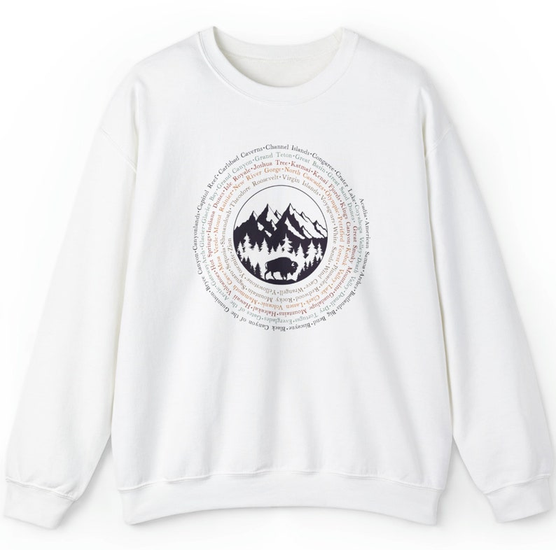 May include: A white crewneck sweatshirt featuring a circular graphic on the chest. The design depicts a black silhouette of a bison standing in front of a forest and mountain range, surrounded by a ring of text listing various national parks.