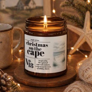 May include: A lit candle in a brown glass jar with a white label featuring the text "maple & hen christmas on the cape No. 18" and a watercolor illustration of a beach scene with tall grass, set on a round wooden coaster with a warm glow.