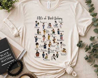 May include: Cream-colored t-shirt featuring the text "ABCs of Black History" with illustrations of notable figures. The design includes various cartoon figures and names, celebrating Black history. The shirt is styled with a knot at the bottom.