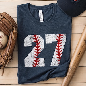 May include: Navy blue t-shirt with a distressed white baseball design featuring the number 27 and red stitching details. A baseball glove, baseball, baseball bat, and baseball cap are also in the image.