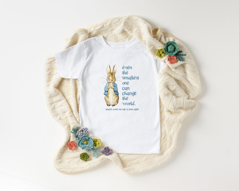 May include: White toddler t-shirt featuring a watercolor illustration of Peter Rabbit wearing a blue jacket. The shirt has the quote "even the smallest one can change the world" in teal text. The quote is attributed to Beatrix Potter.