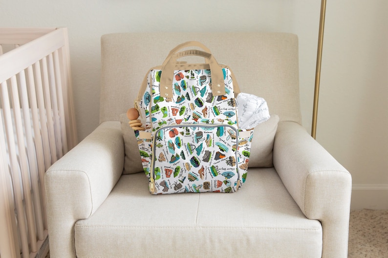 Puede incluir: Un bolso para pa&ntilde;ales blanco con un colorido estampado de varias ilustraciones tem&aacute;ticas al aire libre, incluyendo tiendas de campa&ntilde;a y coches. El bolso tiene detalles en beige, un asa superior y bolsillos laterales. Se ve un pa&ntilde;o blanco y un juguete de madera.