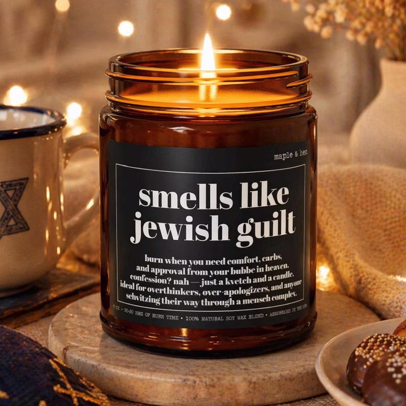 May include: A lit candle in an amber glass jar with a black label featuring the white text: smells like jewish guilt, burn when you need comfort, carbs, and approval from your bubbe in heaven. confession? nah &mdash; just a kvetch and a candle.