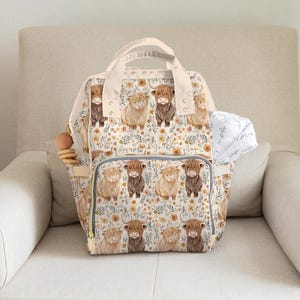 May include: A cream-colored diaper bag with a repeating pattern of Highland cows and floral accents. The bag has multiple pockets, a top handle, and a zipper closure. A wooden teething ring hangs from the side.
