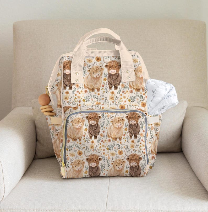 May include: A cream-colored diaper bag with a repeating pattern of Highland cows and floral accents. The bag has multiple pockets, a top handle, and a zipper closure. A wooden teething ring hangs from the side.