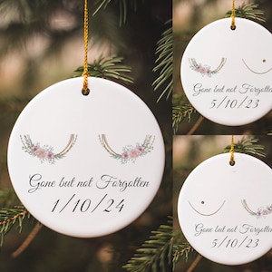 May include: Three white ceramic ornaments with gold string hangers. Each ornament features a stylized breast cancer awareness ribbon with pink flowers and the text "Gone but not Forgotten" with a date.