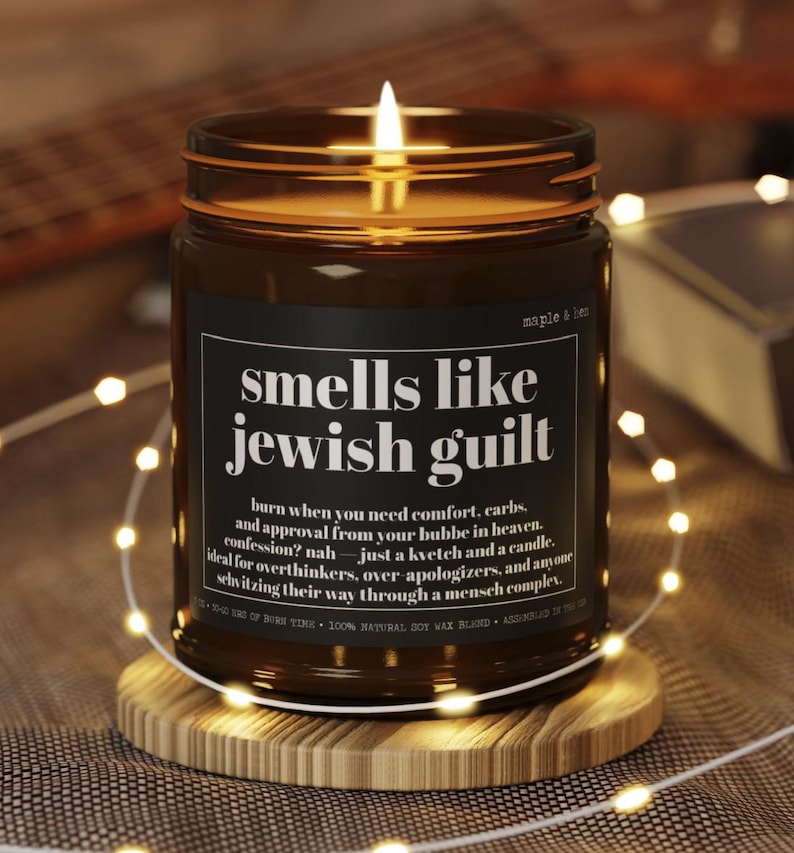 May include: A lit candle in a brown glass jar with a black label featuring the white text: smells like jewish guilt, burn when you need comfort, carbs, and approval from your bubbe in heaven. confession? nah &mdash; just a kvetch and a candle.