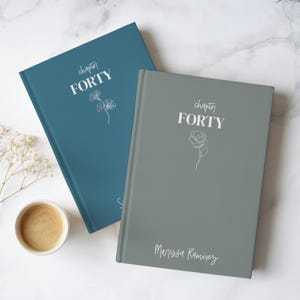 Peut inclure: Deux journaux à couverture rigide, un bleu sarcelle et un gris, sont présentés sur une surface en marbre blanc. Le journal bleu sarcelle porte le texte "chapter FORTY" et une illustration florale. Le journal gris porte le même texte et une illustration de rose, avec le nom "Marissa Ramirez" en dessous.