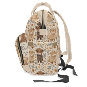May include: A beige backpack with a repeating pattern of Highland cows and floral elements. The backpack has a top handle, adjustable straps, and a zippered main compartment. The design features light brown, dark brown, and yellow colors.