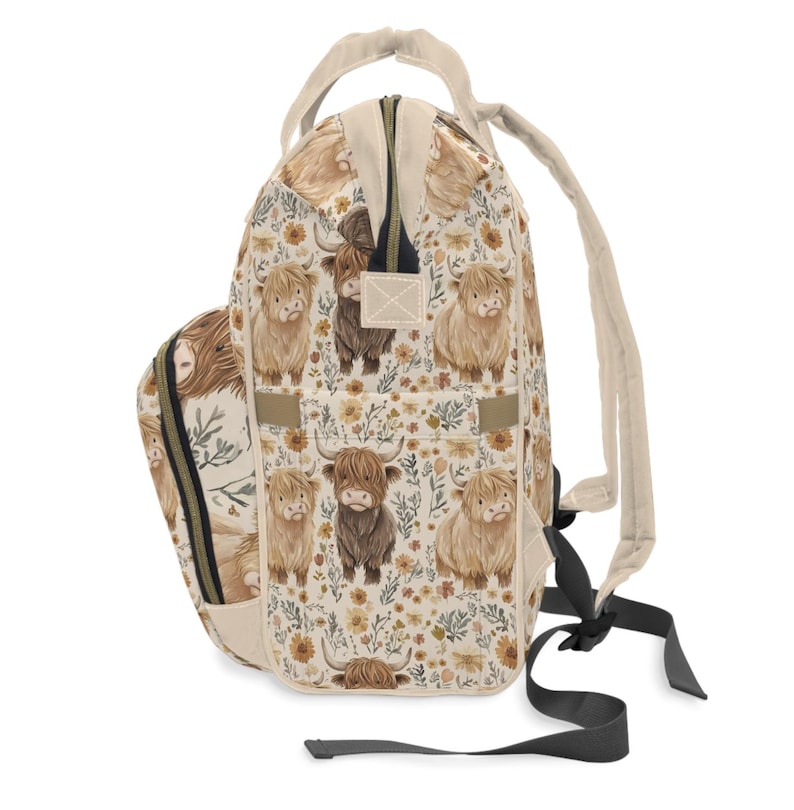 May include: A beige backpack with a repeating pattern of Highland cows and floral elements. The backpack has a top handle, adjustable straps, and a zippered main compartment. The design features light brown, dark brown, and yellow colors.