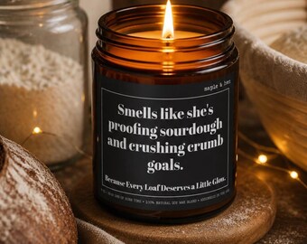 May include: A lit candle in an amber glass jar with a black label featuring the white text: Smells like she's proofing sourdough and crushing crumb goals. Because Every Loaf Deserves a Little Glow. The candle sits on a round wooden coaster.