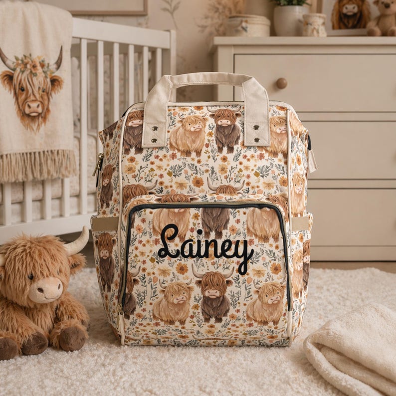 May include: A floral-patterned diaper bag featuring illustrations of Highland cows. The bag has multiple pockets and a top handle. The name "Lainey" is written in cursive on the front. A stuffed Highland cow and a baby blanket are also in the image.