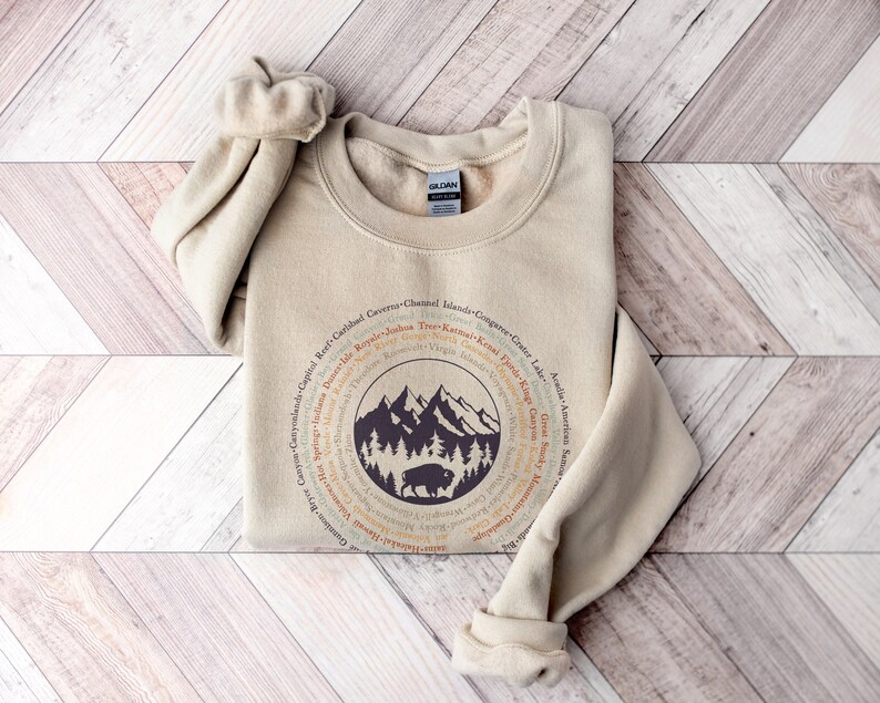 May include: A sand-colored crewneck sweatshirt featuring a circular graphic design. The center displays a dark purple illustration of mountains, pine trees, and a bison. Surrounding the central image are the names of various national parks.