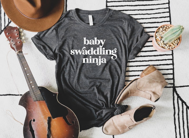 Puede incluir: Camiseta gris oscuro con la frase "baby swaddling ninja" en blanco. La imagen incluye un sombrero marr&oacute;n, una mandolina, botas color beige y un peque&ntilde;o cactus en una maceta rosa, sobre una alfombra.