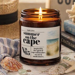 May include: A brown glass jar candle with a lit wick. The white label features the text "maple & hen summer on the cape No. 65" and a watercolor illustration of a beach with sand, grass, and blue water. The candle is surrounded by seashells.