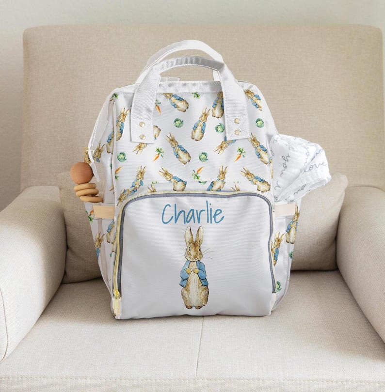 May include: White baby changing bag with a Peter Rabbit design. The bag has a repeating pattern of Peter Rabbit, carrots, and green leaves. The front pocket features the name "Charlie" in blue script and a Peter Rabbit illustration. A wooden teething ring is in the side pocket.