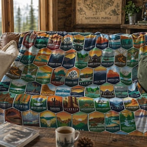 May include: A colorful blanket featuring hexagonal patches with illustrations of various National Parks. The blanket displays images of mountains, lakes, and trees, with park names like "Crater Lake" and "Grand Canyon". The blanket is draped over a brown leather couch.