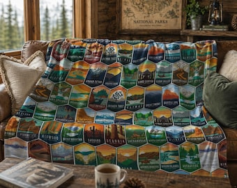 May include: A colorful blanket featuring hexagonal patches with illustrations of various National Parks. The blanket displays images of mountains, lakes, and trees, with park names like "Crater Lake" and "Grand Canyon". The blanket is draped over a brown leather couch.