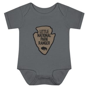 May include: A dark grey short-sleeved baby bodysuit featuring a tan arrowhead-shaped graphic on the chest. The graphic contains the black text "LITTLE NATIONAL PARK RANGER" and a small black silhouette of a bison at the bottom of the arrowhead.