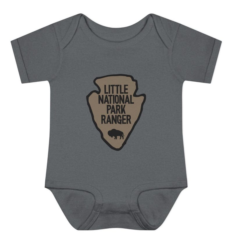 May include: A dark grey short-sleeved baby bodysuit featuring a tan arrowhead-shaped graphic on the chest. The graphic contains the black text "LITTLE NATIONAL PARK RANGER" and a small black silhouette of a bison at the bottom of the arrowhead.