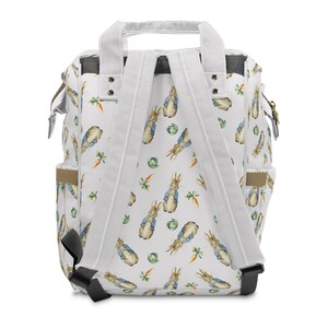May include: White backpack featuring a pattern of Peter Rabbit illustrations, carrots, and cabbage. It has white straps, a top handle, and side pockets. The design is inspired by a classic children's book.