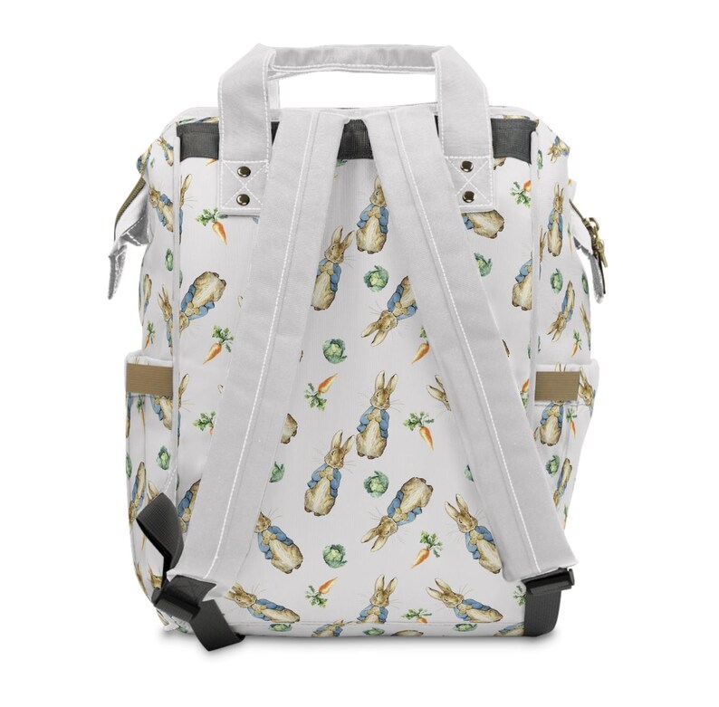 May include: White backpack featuring a pattern of Peter Rabbit illustrations, carrots, and cabbage. It has white straps, a top handle, and side pockets. The design is inspired by a classic children's book.
