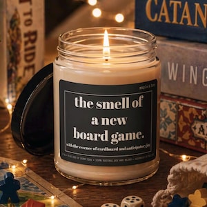 May include: A clear glass jar candle with a lit wick and a black label featuring white text that reads: the smell of a new board game. The label also includes the text: maple & ben, with the essence of cardboard and anticipatory joy.