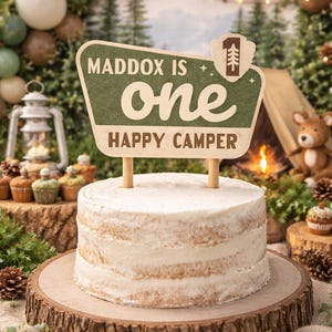 Op de afbeelding: Een gelaagde taart met witte frosting op een houten taartstandaard. Een bord met de tekst "Maddox is one Happy Camper" staat bovenop de taart. De achtergrond heeft een campingthema met ballonnen, een tent en cupcakes.